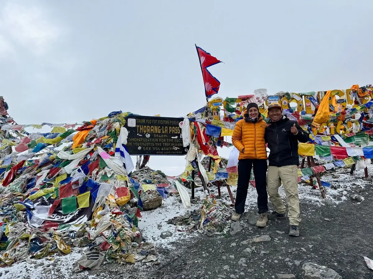 Two trekkers beside the Thorong La Pass sign surrounded by prayer flags and snow